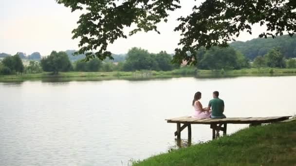 Hermosa pareja de youn sentarse en el muelle cerca del lago. Ambiente romántico — Vídeos de Stock