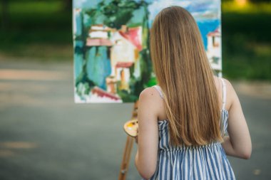 Taburede oturan güzel sarışın kız ve parkta boyalar ve bir spatula ile bir palet kullanarak bir resim çizer