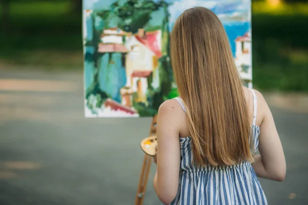 Schöne blonde Haare Mädchen sitzt auf einem Hocker und zeichnet ein Bild im Park mit einer Palette mit Farben und einem Spachtel — Stockfoto