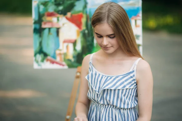 Hermosa chica de pelo rubio sentada en el taburete y dibuja un cuadro en el parque usando una paleta con pinturas y una espátula —  Fotos de Stock
