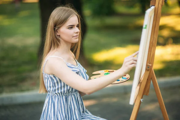 Krásné blond vlasy dívka sedí na stoličce a nakreslí obrázek v parku pomocí palety barvy a stěrky — Stock fotografie