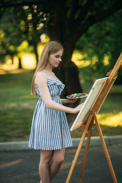 Beautiful girl draws a picture in the park using a palette with paints and a spatula. Easel and canvas with a picture — Stock Photo, Image