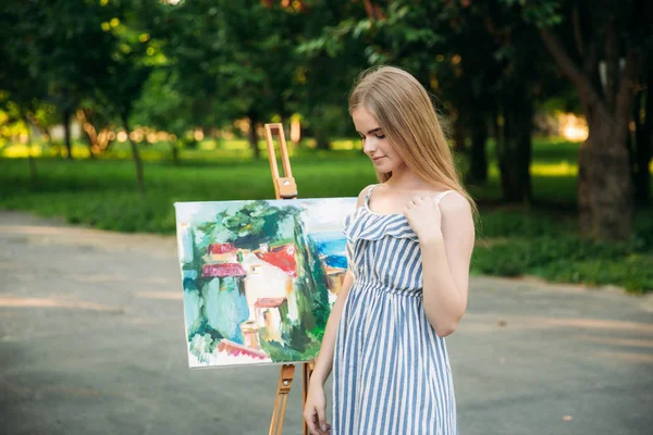 Hermosa artista chica dibuja un cuadro en el parque utilizando una paleta con pinturas y una espátula. caballete y lienzo con una imagen —  Fotos de Stock