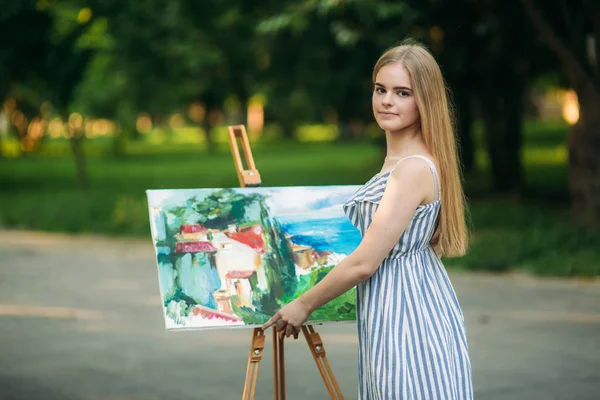 Hermosa artista chica dibuja un cuadro en el parque utilizando una paleta con pinturas y una espátula. caballete y lienzo con una imagen — Foto de Stock