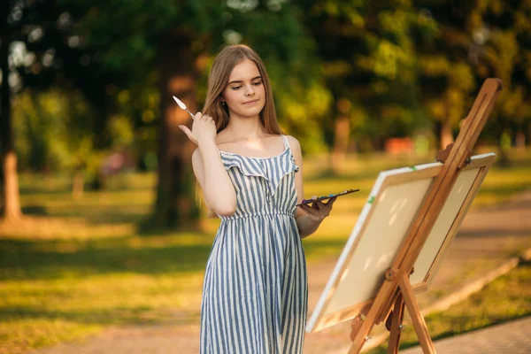 Menina de cabelo loiro no vestido desenhando um quadro no parque — Fotografia de Stock