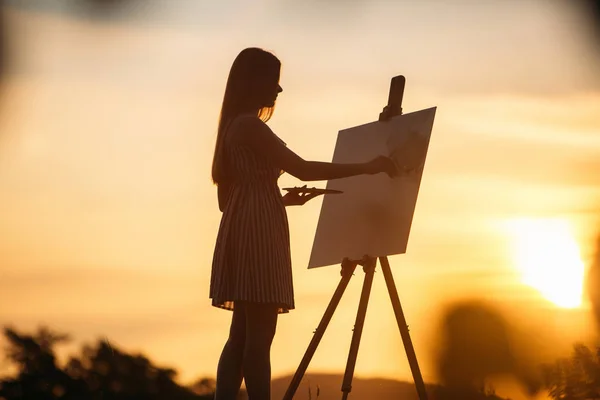 Silhouette of a blonde girl artist. Lady paints a painting on the canvas with the help of paints. A wooden easel keeps the picture. Summer is a sunny day, sunset