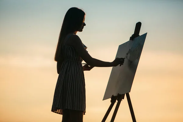 Silhouette di una ragazza bionda artista. Lady dipinge un dipinto sulla tela con l'aiuto di vernici. Un cavalletto di legno mantiene il quadro. L'estate è una giornata di sole, tramonto — Foto Stock