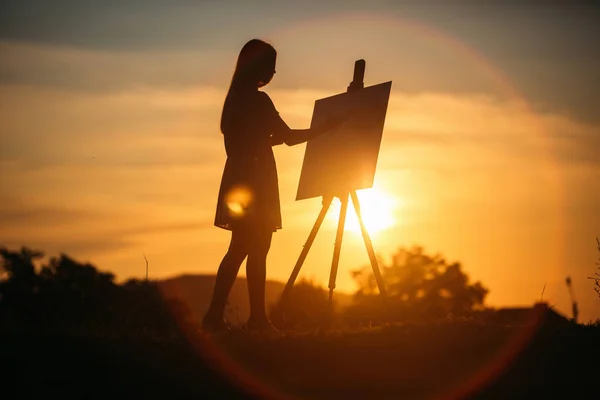 Silhouette of a blonde girl artist. Lady paints a painting on the canvas with the help of paints. A wooden easel keeps the picture. Summer is a sunny day, sunset