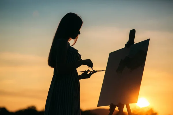 Silhueta de uma artista loira. Lady pinta uma pintura na tela com a ajuda de tintas. Um cavalete de madeira mantém a imagem. O verão é um dia ensolarado, pôr do sol — Fotografia de Stock