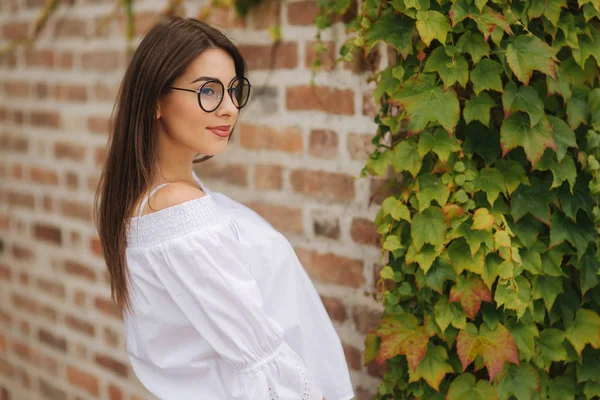 Boldog női napszemüveg portréja. Lány öltözött egy fehér inget áll közel a fal, benőtt a borostyán — Stock Fotó