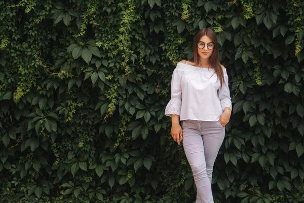 Girl girl smile to camera. Female is dressed in a white shirt is standing near a wall overgrown with ivy — Stock Photo, Image