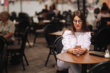 Kafe ve manifatura akıllı telefon üzerinde oturan iş kız. o bir beyaz gömlek ve sunglasess giymiş