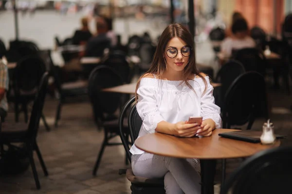 商业女孩坐在咖啡馆里, 用智能手机发短信。她穿着一件白衬衣和 sunglasess。 — 图库照片