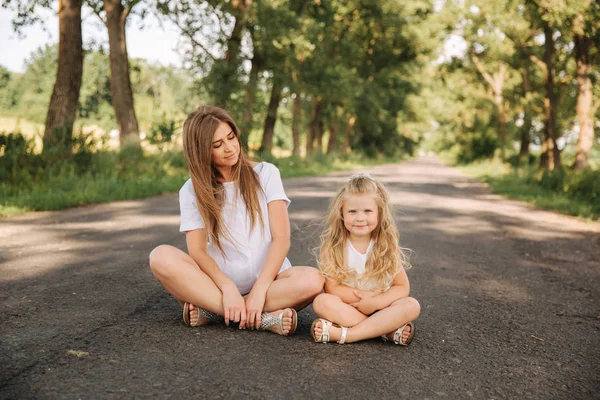 Attraktive Mutter und blonde Haare Tochter sitzt auf der Straße in der Nähe der großen Gasse. sie lächeln und schauen zur Natur. Frontansicht — Stockfoto