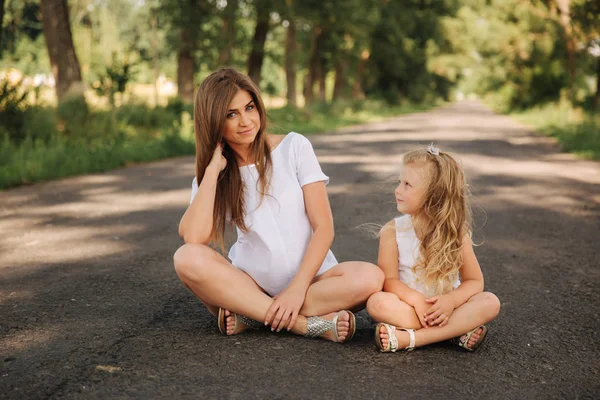 Attractive Mom and blonde hair daughter sits on road near big alley. They smile and look to natune. Front view