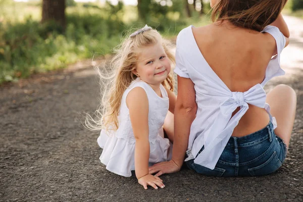 Attractive Mom and blonde hair daughter sits on road near big alley. They smile and look to natune — Stock Photo, Image