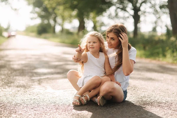 Attractive Mom and blonde hair daughter sits on road near big alley. They smile and look to natune — Stock Photo, Image