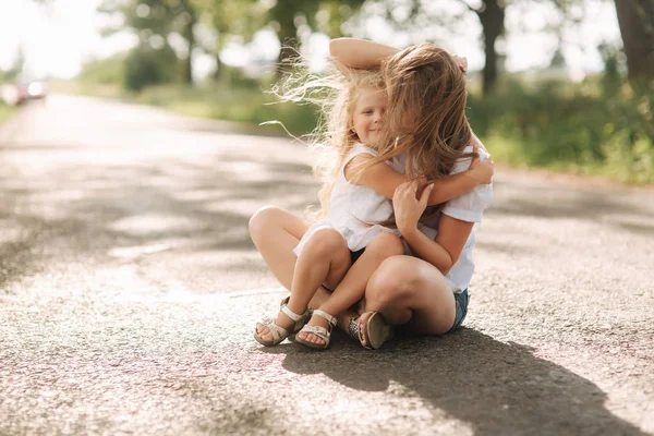 Attractive Mom and blonde hair daughter sits on road near big alley. They smile and look to natune — Stock Photo, Image