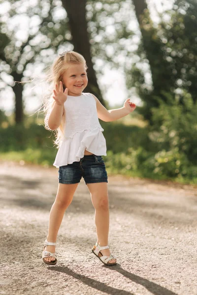 Een vrolijke schattig klein meisje met blond haar dansen en springt via het steegje op een warme zomerdag — Stockfoto