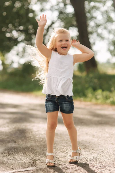 Una niña alegre y adorable con el pelo rubio baila y salta por el callejón en un cálido día de verano — Foto de Stock