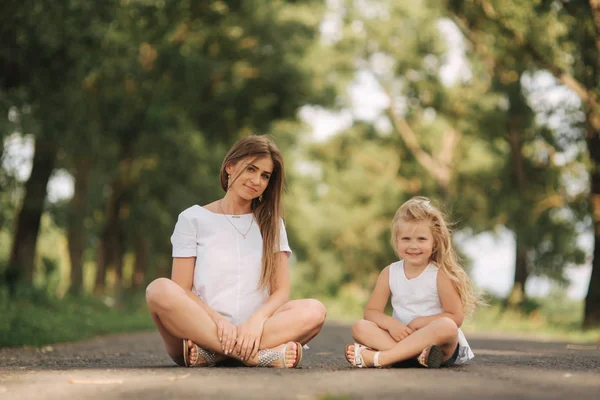 Attractive Mom and blonde hair daughter sits on road near big alley. They smile and look to natune. Front view — Stock Photo, Image