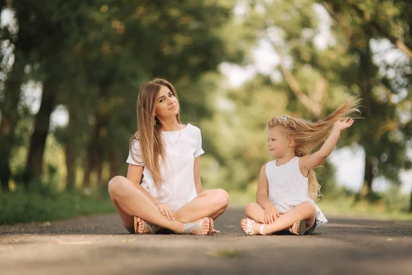 Atraktivní máma a dcera sedí na silnici poblíž velké alej blond vlasy. Úsměv a podívejte se na natune. Pohled zepředu — Stock fotografie