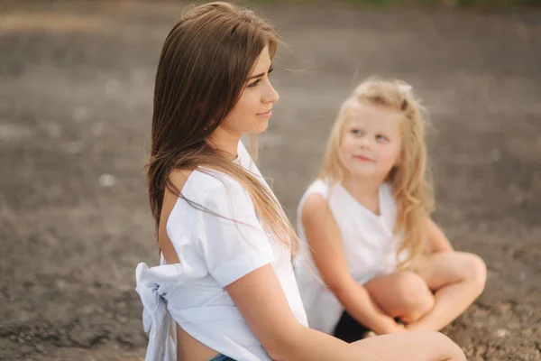 Attractive Mom and blonde hair daughter sits on road near big alley. They smile and look to natune — Stock Photo, Image