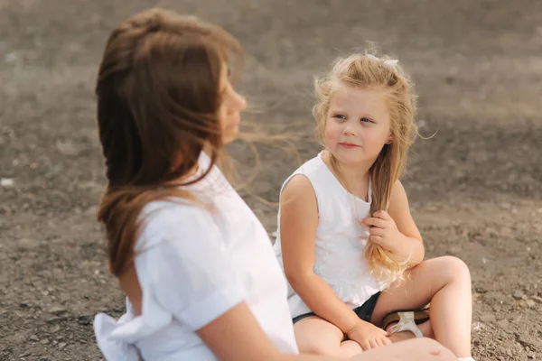 Attractive Mom and blonde hair daughter sits on road near big alley. They smile and look to natune