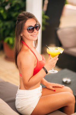 Renkli bir kokteyl beach club bar bir kabin üzerinde oturan içme mayo giyen güzel genç kadın. Çarpıcı kız onun içeceğinizi üzerinde yaz saati