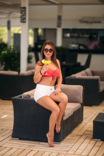 Mulher bonita vestindo maiô bebendo um coquetel colorido sentado em uma cabine do bar do clube de praia. Menina impressionante desfrutando de sua bebida na hora de verão — Fotografia de Stock