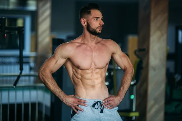 Jovem atleta posando com um tronco para fotografia em um fundo de ginásio. Fisiculturista, atleta com músculos bombeados — Fotografia de Stock