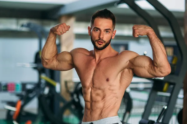 Jovem atleta posando com um tronco para fotografia em um fundo de ginásio. Fisiculturista, atleta com músculos bombeados — Fotografia de Stock