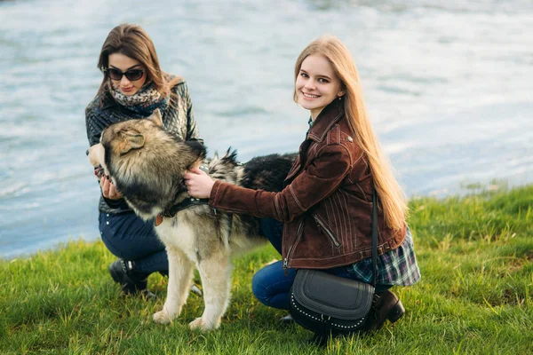 Deux sœurs marchent avec un chien husky devant la rivière. Brunettle et blonde fille et caresse un chien — Photo