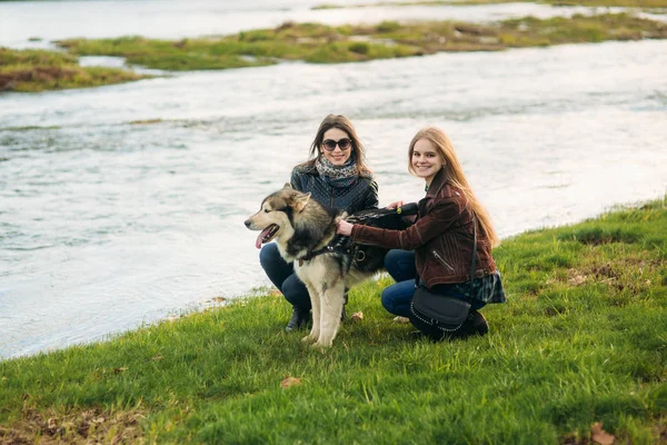 Zwei Schwestern spazieren mit Husky-Hund vor dem Fluss. brünett und blonde Mädchen und streicheln einen Hund — Stockfoto