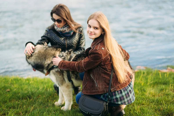 Due sorelle camminano con il cane husky di fronte al fiume. Bruna e bionda ragazza e accarezzare un cane — Foto Stock