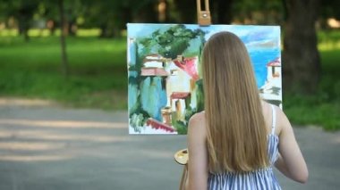 Güzel kız parkta oturan ve boyalar ve bir spatula ile bir palet kullanarak resim çizer.