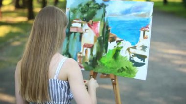 Güzel kız parkta oturan ve boyalar ve bir spatula ile bir palet kullanarak resim çizer.