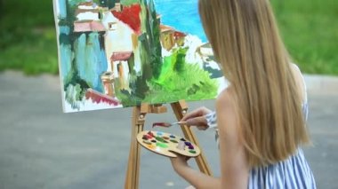 Güzel kız parkta oturan ve boyalar ve bir spatula ile bir palet kullanarak resim çizer.