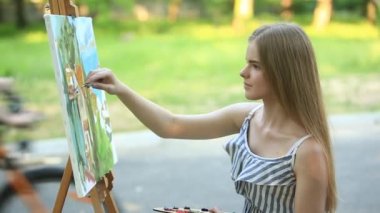 Güzel kız parkta oturan ve boyalar ve bir spatula ile bir palet kullanarak bir resim çizer. yaz