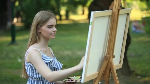 Красива дівчина сидить в парку і малює картину, використовуючи палітру з фарбами і шпателем . — стокове відео