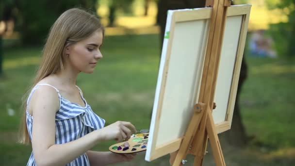 Hermosa chica sentada en el parque y dibuja un cuadro usando una paleta con pinturas y una espátula . — Vídeos de Stock