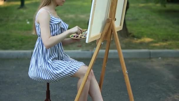 Bella ragazza seduta nel parco e disegna un disegno utilizzando una tavolozza con vernici e una spatola . — Video Stock