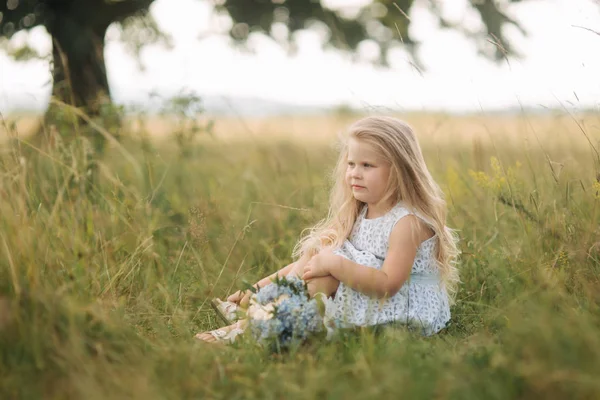 Klein meisje in hemelsblauw jurk met boeket zit in veld voor grote boom. Kind lachen en plezier — Stockfoto