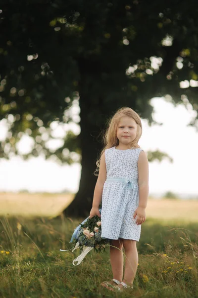 Niña en vestido azul cielo con ramo de pie en el campo delante de un gran árbol. Niño sonrisa y divertirse — Foto de Stock