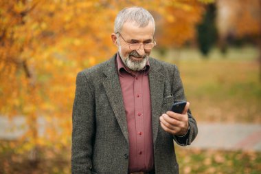 Giyen bir yakışıklı elderny adam bir telefonu kullanarak gözlük. Sonbaharda parkta yürümek