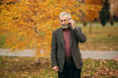Giyen bir yakışıklı elderny adam bir telefonu kullanarak gözlük. Sonbaharda parkta yürümek