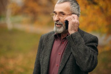 Giyen bir yakışıklı elderny adam bir telefonu kullanarak gözlük. Sonbaharda parkta yürümek