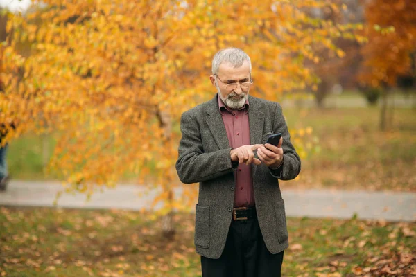 Un bell'uomo anziano con gli occhiali al telefono. Passeggiata nel parco in autunno — Foto Stock