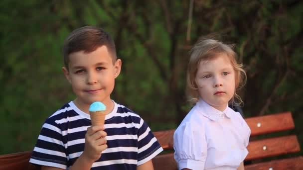 Niño comiendo un helado y sentado en el banco mientras que la chica está buscando — Vídeos de Stock