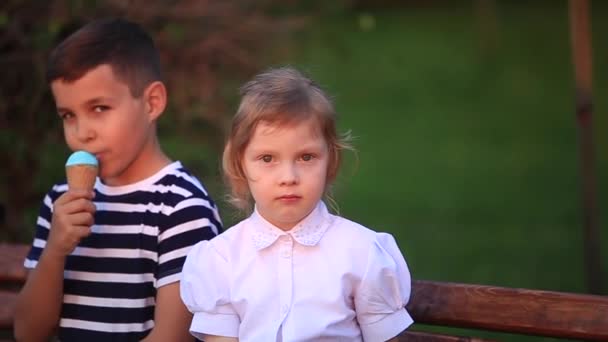 Chłopiec, jedzenie lodów i siedząc na ławce, a dziewczyna szuka — Wideo stockowe